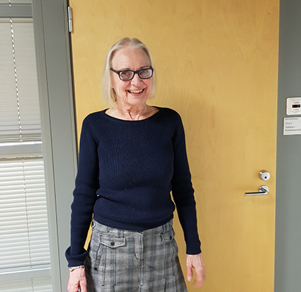Helena Vaittinen seisoo sisällä toimiston oven edessä ja hymyilee. Hänellä on tummansininen paita ja vaalean harmaa hame.