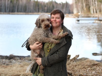 Reetta seisoo järven rannalla ja pitää sylissään koiraa.
