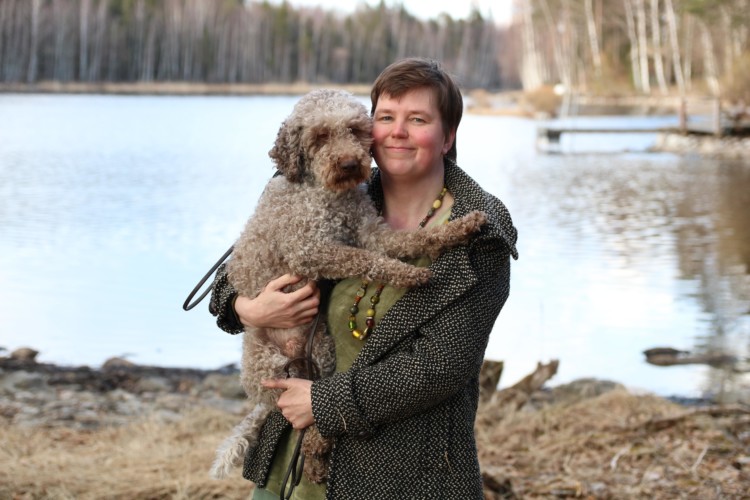 Reetta seisoo järven rannalla ja pitää sylissään koiraa.