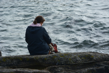 Nainen istuu rantakalliolla ja katsoo alaviistoon.