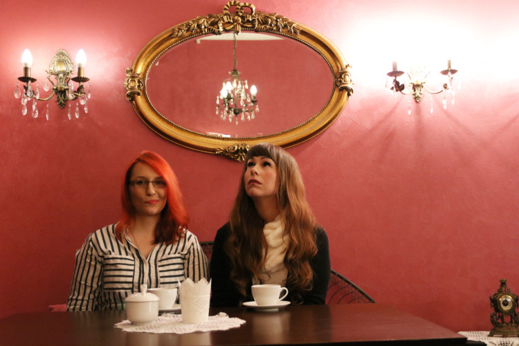 Kaksi naista istuvat kahvilapöydän ääressä. Takana punainen seinä, jolla juhlava peili ja kyntteliköt.