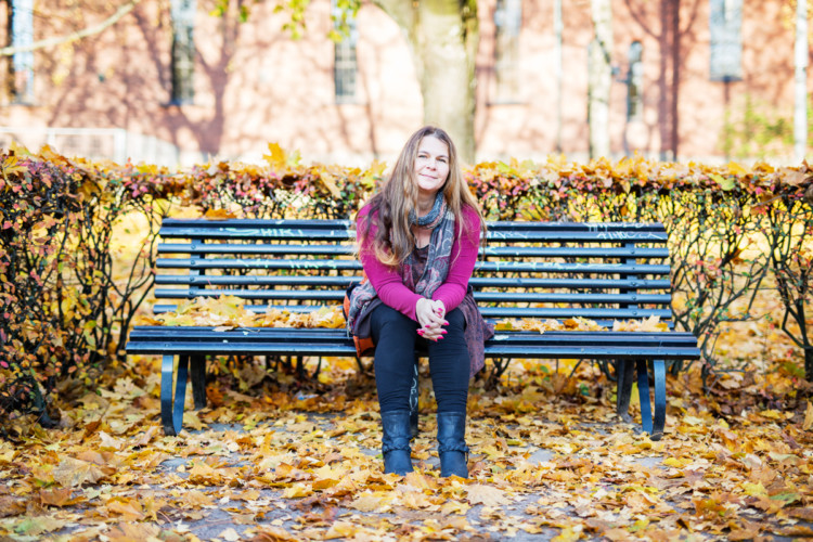 Hymyilevä nainen istuu penkillä, maassa ja penkillä on paljon tippuneita syyslehtiä.