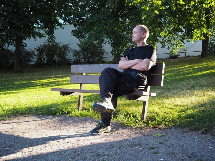 Jari istuu puistonpenkillä ja katsoo poispäin kamerasta. Taustalla nurmikkoa ja puita.