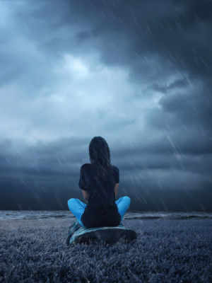 Ihminen istuu sateessa vesistön rannalla. Taivas veden yläpuolella on hyvin tumma ja synkkä.