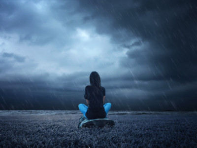 Ihminen istuu sateessa vesistön rannalla. Taivas veden yläpuolella on hyvin tumma ja synkkä.