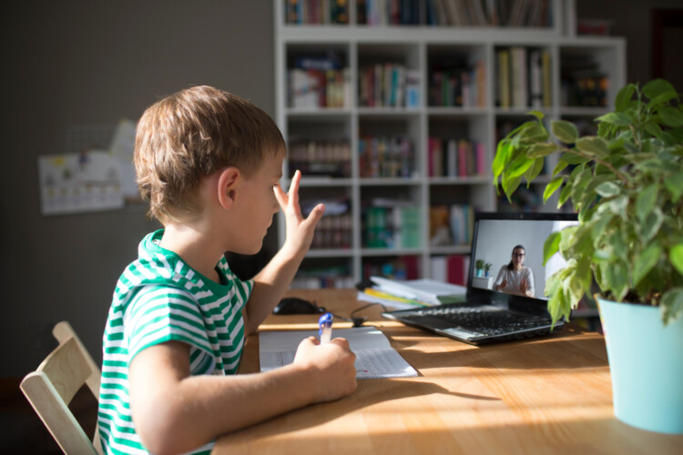 Poikamaisen oloinen lapsi etäkoulussa tietokoneen ääressä. Poika kirjoittaa vihkoon samalla kun ruudulla näkyy opettaja puhumassa.