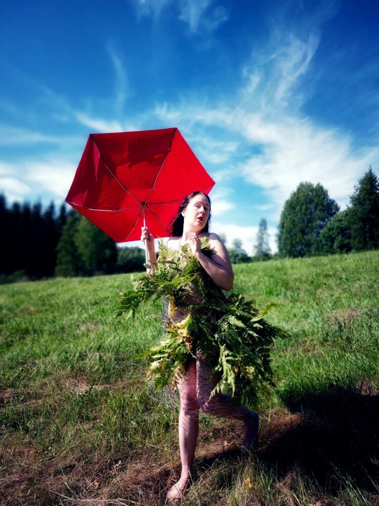 Elina alias Inkeri Routakorpi kesämaisemassa. Hänellä on väärinpäin kääntynyt sateenvarjo kädessä ja hän on pukeutunut kasvien lehtiin.