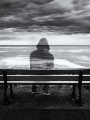 Mustavalkokuvassa penkillä istuu huppupäinen läpinäkyvä ihmishahmo, joka katsoo tuuliselle merelle. Synkkä pilvinen taivas.