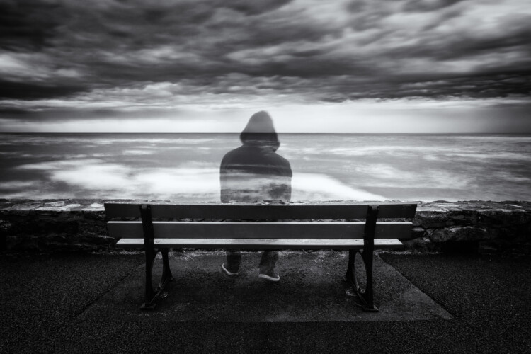 Mustavalkokuvassa penkillä istuu huppupäinen läpinäkyvä ihmishahmo, joka katsoo tuuliselle merelle. Synkkä pilvinen taivas.