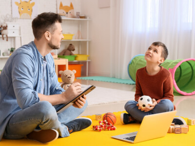 Mies istuu lattialla pienen pojan kanssa, jonka katse on suunnattu kattoon. Pojalla on kädessä pallo, lattialla rakennuspalikoita ja leluja sekä kannettava tietokone.