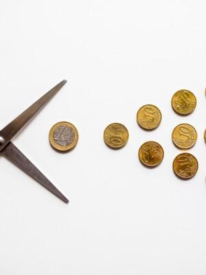 Avatut sakset ja kolmion muotoisesti asetetut 10- ja 20 eurosentin kolikot sekä yksi euron kolikko vaalealla taustalla.