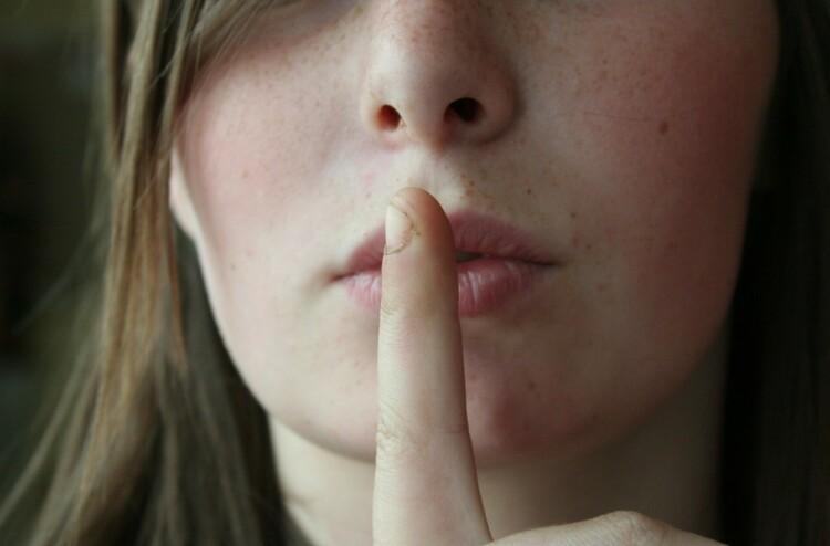 Lähikuva henkilöstä, joka pitää sormeaan suun edessä hiljaisuuden merkiksi.