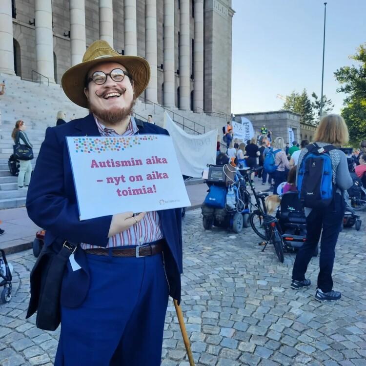 Orjo Pättiniemi eduskuntatalon edessä kädessä kyltti Autismin aika - nyt on aika toimia!