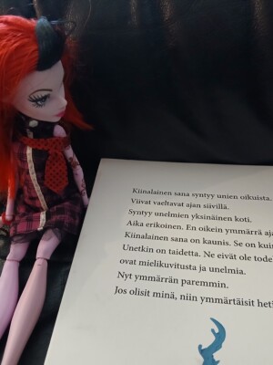 Kuvassa punatukkainen Monster High -nukke istuu kirjan vieressä ja "lukee" kirjaa.