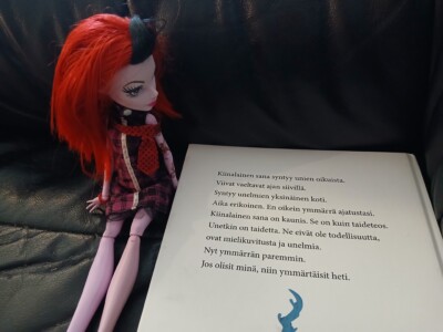 Kuvassa punatukkainen Monster High -nukke istuu kirjan vieressä ja "lukee" kirjaa.