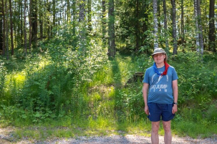 Mies lippiksellä ja partiohuivilla seisoo kesän vehreydessä metsän laidalla.