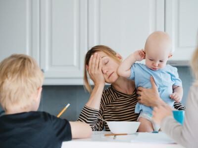 Väsynyt äiti ruokapöydän ääressä. Hän pitää sylissään taaperoa ja pöydän ääressä vähän isompia lapsia tekemässä läksyjä.