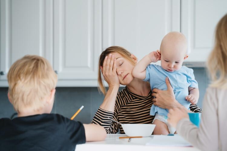Väsynyt äiti ruokapöydän ääressä. Hän pitää sylissään taaperoa ja pöydän ääressä vähän isompia lapsia tekemässä läksyjä.