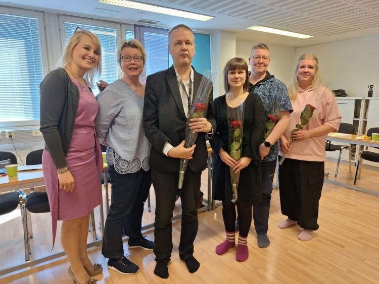 Kuusi henkilöä ryhmäkuvassa, osalla kädessä punainen ruusu.
