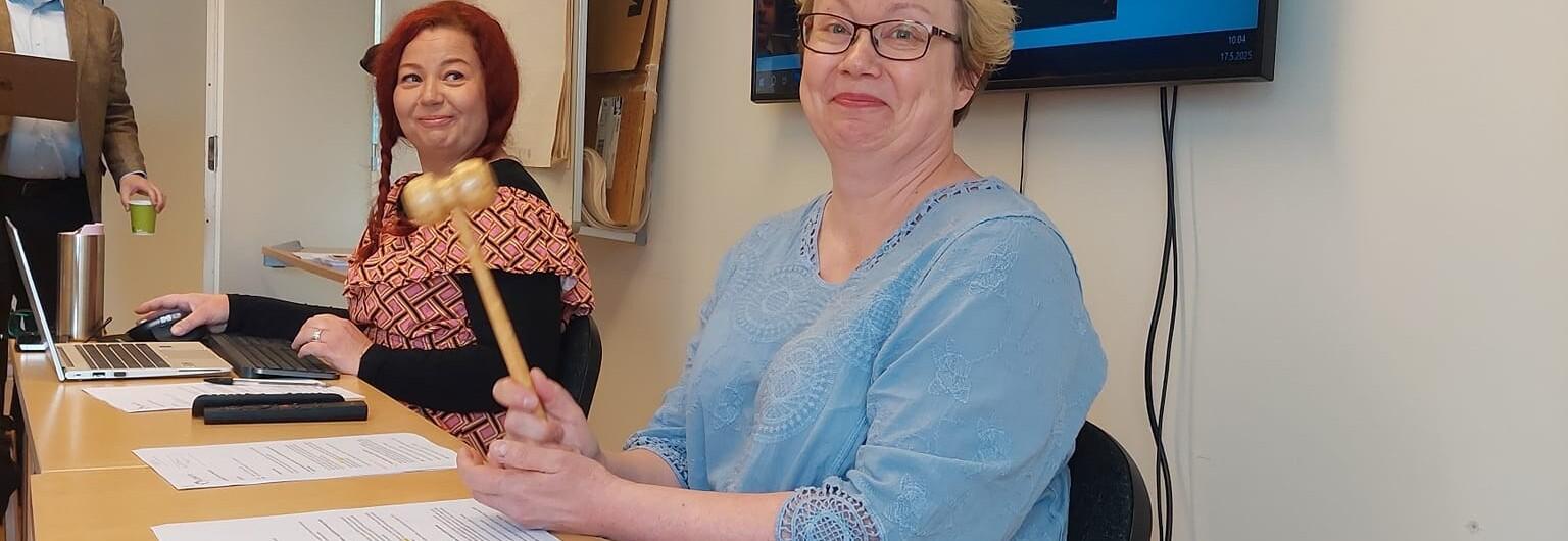 Etualalla Autismiliiton hallituksen puheenjohtaja Petra Hietala-Rasku heiluttaa puheenjohtajan vasaraa, vieressä istuu Autismiliiton hallintosihteeri Johanna Eestilä ja taustalla kävelee Teemu Muukkonen.
