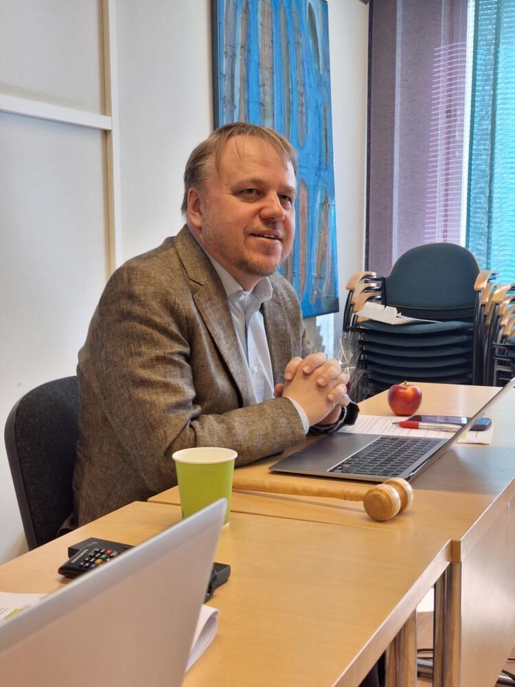 Teemu Muukkonen istuu tietokoneen ääressä, vieressä mm. puheenjohtajan vasara ja omena.