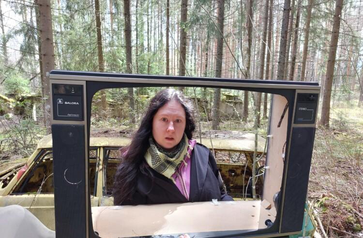 Nainen on metsässä ja pitää edessään vanhaa television kehystä, taustalla autoromu.