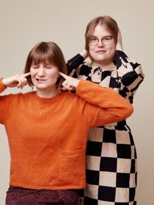 Studiokuva, jossa oranssipaitainen Heli Rantala istuu. Hänen vieressään seisoo ruutumekkoon pukeutunut Netta Lehtola. Molemmat painavat käsillä korviaan.