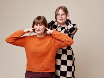 Studiokuva, jossa oranssipaitainen Heli Rantala istuu. Hänen vieressään seisoo ruutumekkoon pukeutunut Netta Lehtola. Molemmat painavat käsillä korviaan.