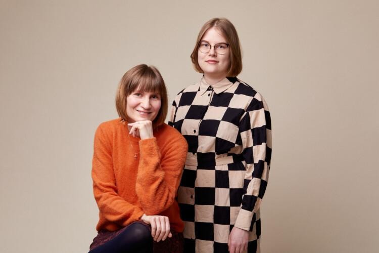 Studiokuva, jossa oranssipaitainen Heli Rantala istuu. Hänen vieressään seisoo ruutumekkoon pukeutunut Netta Lehtola.