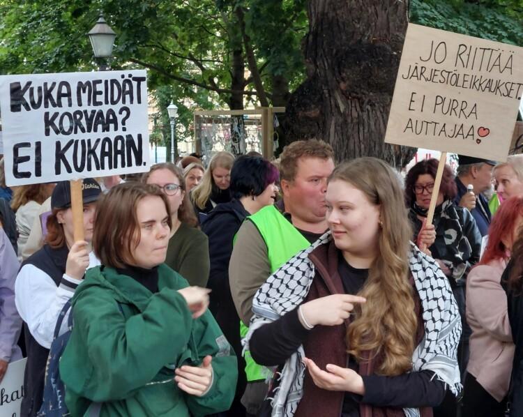 Ihmisiä mielenosoituksessa. Etualalla kaksi henkilöä viittomassa. Kylteissä lukee Kuka meidät korvaa? -Ei kukaan. Jo riittää järjestöleikkaukset. Ei purra auttajaa.