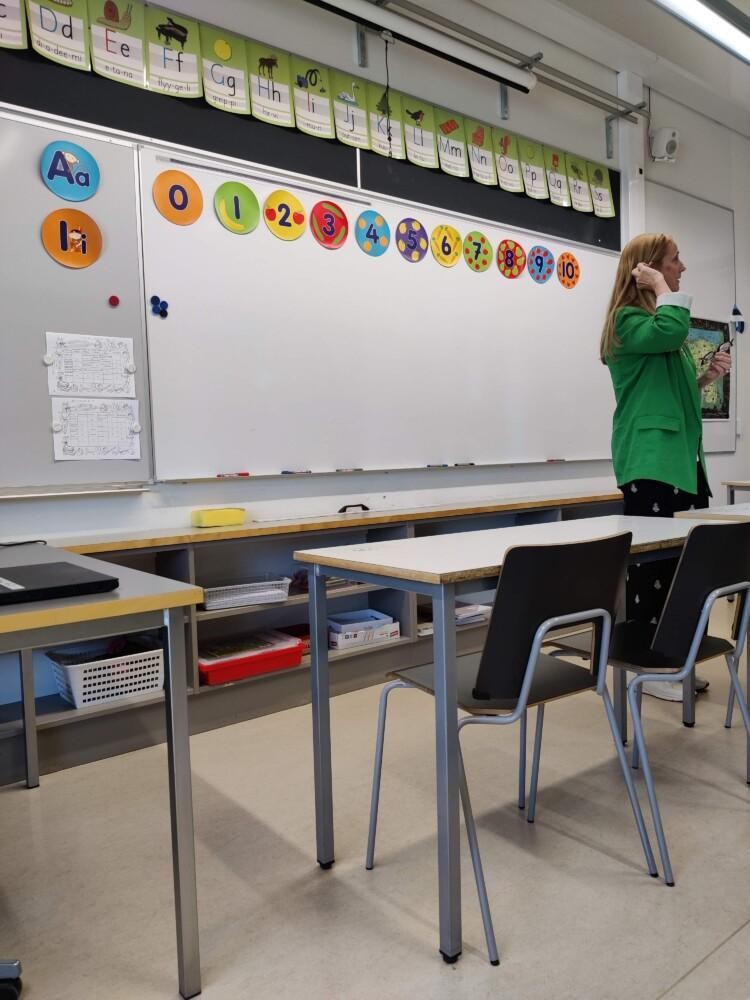 Koululuokka, jossa on valkotaulu, värikkäitä kirjaimia ja numeroita, kuvan laidassa opettaja.