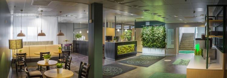 Hotellin aula, jossa näkyy viherseinä, sohva, tuoleja, pöytiä, kahvipiste, vastaanottopöytä ja portaat. Lattialla mattoja.