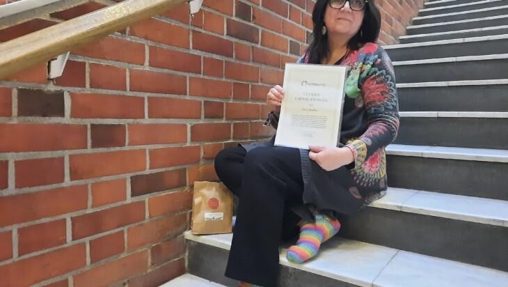 Maria Kuoksa istuu portailla ja hänellä on kädessään vuoden vapaaehtoisen kunniakirja.