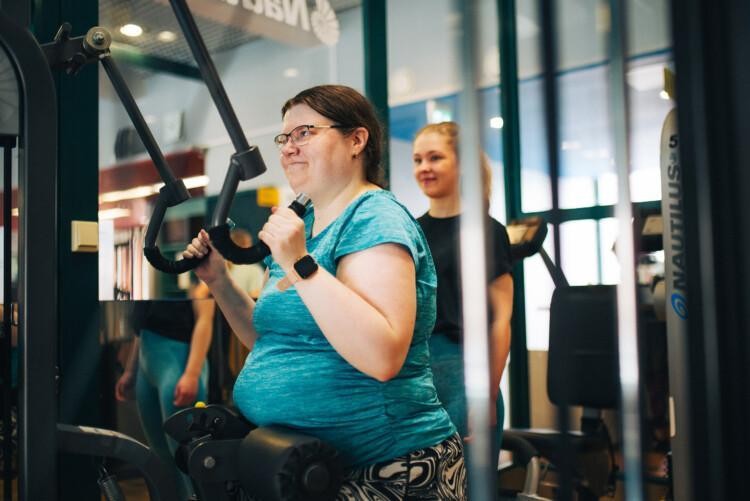 Maija Juhola treenaa kuntosalin laitteella, Laura Rautio taustalla seuraamassa.
