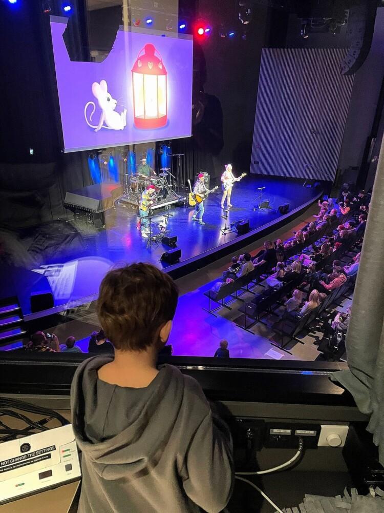 Lapsi seuraa konserttia lavan yläpuolelta erillisessä tilassa, lavalla soittamassa bändi, valkokankaalla iso kuva kiirestä ja lyhdystä, katsomossa yleisöä.