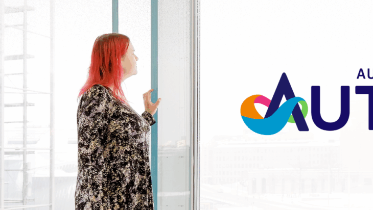 Punapäinen nainen katsoo lasiseinäikkunasta ulos, taustalla utuisena Eduskuntatalo ja Autismi-lehden logo