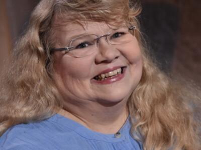 Kuvassa hymyilevä Annikka Suoninen. Hänellä on sininen paita, vaalea, pitkä, hieman kihara tukka ja lismälasit.