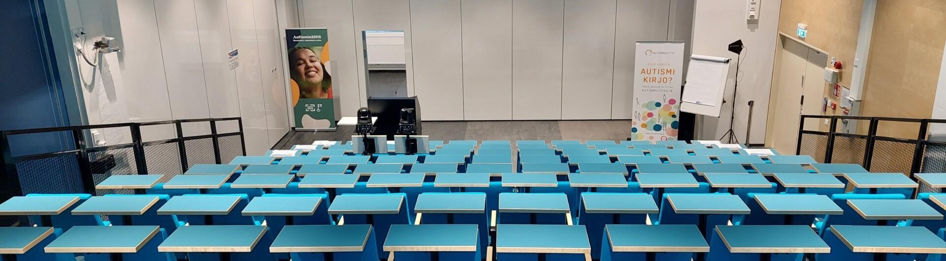 Tyhjä auditoriosali, jossa turkoosit pöydät ja penkit.