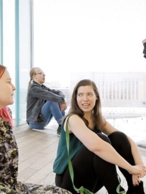 Kaksi naista istuvat jutellen lattialla ja kaksi miestä keskustelevat lasi-ikkunan luona Oodi-kirjastossa, taustalla eduskuntatalo.