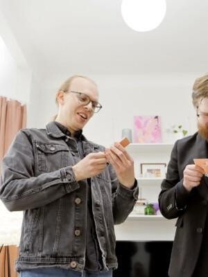 Kaksi miestä seisovat ja katsovat hymyillen käsissään olevia pelikortteja.