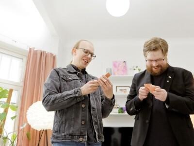 Kaksi miestä seisovat ja katsovat hymyillen käsissään olevia pelikortteja.