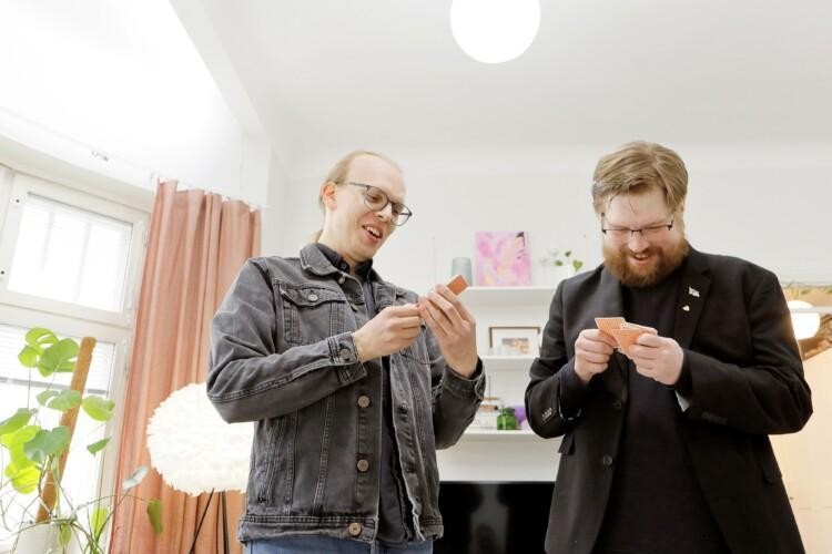Kaksi miestä seisovat ja katsovat hymyillen käsissään olevia pelikortteja.