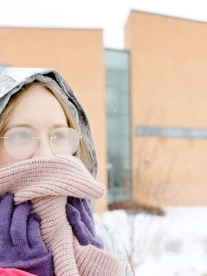 Talvisesti pukeutunut henkilö on peittänyt suunsa huivilla. Hänen silmälasinsa ovat huurussa.